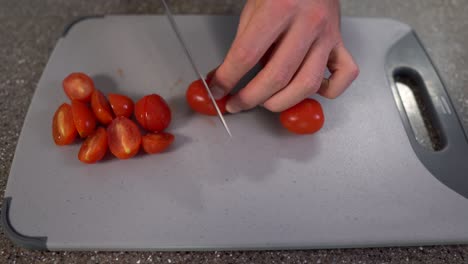 Man-hands-cutting-the-cherry-tomatoes-into-half-pieces-with-chefs-knife-on-grey-chopping-board-on-the-table,-top-view