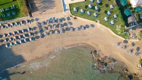 Aerial-view-over-the-calm-sea-water-reaching-the-beach-with-shacks-arranged-systematically-at-tourist-destination-in-Greek-island