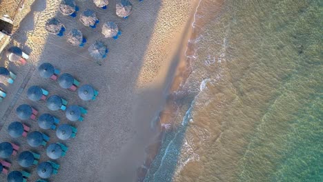 Luftaufnahme-Von-Oben-Nach-Unten-über-Wellen,-Die-Am-Strand-Mit-Leeren-Sonnenliegen-Mit-Blauen-Sonnenschirmen-Zusammenbrechen