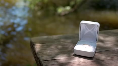 Gold-engagement-ring-in-white-box-sits-on-wooden-table-by-river,-dolly-push