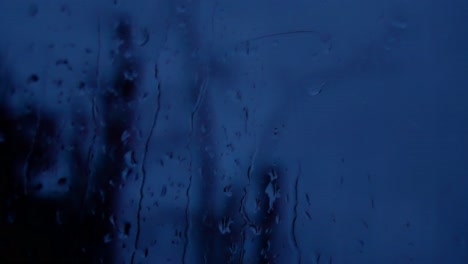Rainy-storm-droplets-at-night,-moody-weather-fall-down-window-glass-closeup-bokeh