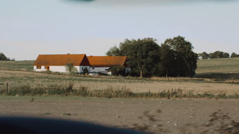 Ansichten-Auf-Einem-Roadtrip-In-Südschweden-Urlaub-Auf-Einem-Kleinen-Bauernhof-Mit-Klassischen-Weißen-Gebäuden-Und-Orangefarbenen-Dächern-Umgeben-Von-Kulturen-Ackerland-Agrarkultur-Aus-Dem-Innenraum-Eines-Autos-Auf-Einer-Straße