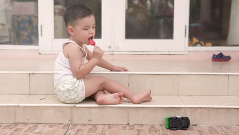 Slow-motion-video-of-a-handsome-Asian-kid-from-the-side-eating-a-posicle-ice-cream-sitting-on-the-stairs-of-a-house-in-summer