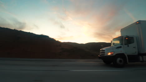 Trucks-Travelling-In-The-Highways-Along-With-The-Other-Vehicles-Passing-By-With-Beautiful-Mountains-During-Sunset---Wide-Shot