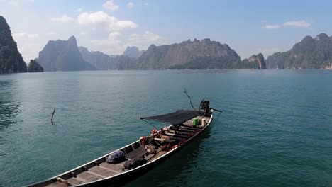 Vista-De-Cerca-De-Un-Bote-De-Madera-Flotando-En-El-Lago-Azul-Turquesa-En-Khao-Sok,-Tailandia-Con-Hermosas-Formaciones-Rocosas-En-El-Fondo---Lapso-De-Tiempo