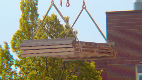 Close-up-of-a-crane-slowly-lifting-and-moving-pieces-of-construction-site-wood-plywood-building-blocks-for-a-building-project-with-string-ropes-and-chains-from-right-to-left-in-frame-on-a-spring-day