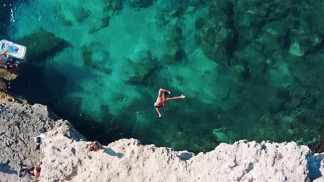 Slow-motion-aerial-top-down-shot-of-man-jumping-from-a-cliff-into-clear-turquoise-water-in-Mallorca,-Spain---Balearic-Island-Holiday