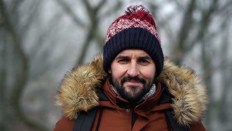 Bearded-Caucasian-male-in-warm-clothes-looks-at-camera-and-smiles-in-the-forest-during-cold-winter-day,-SLOW-MOTION