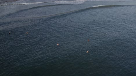 Surfers-waiting-on-surfboards-behind-wave-breaks,-aerial-view-circling-above