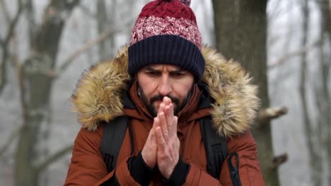 Bearded-Caucasian-male-standing-in-forest-and-warming-up-his-hands-with-breath-during-cold-winter-day,-SLOW-MOTION