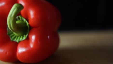 Red-bell-pepper-rolling-on-wooden-chopping-board-on-black-background,-SLOW-MOTION