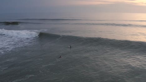 Surfers-paddling-over-waves-in-coastal-waters,-sunset-aerial-view