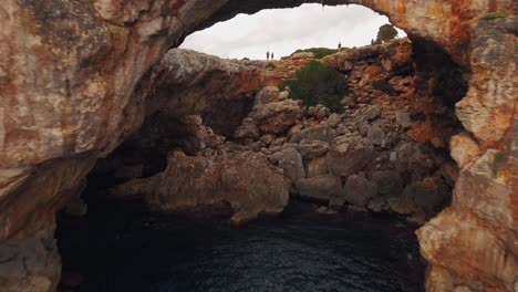 Touristen,-Die-Auf-Einer-Klippe-Stehen---Fliegen-Mit-Einer-Drohne-Durch-Eine-Höhle-An-Der-Wunderschönen-Küste-Von-Mallorca,-Spanien---4k