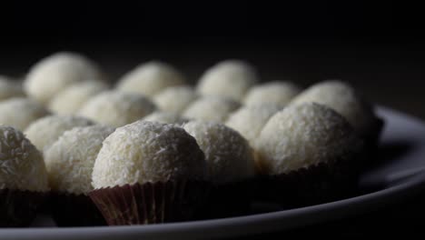 Sweet-white-balls-covered-in-dried-coconut-sitting-in-little-brown-paper-liners-on-white-ceramic-plate,-PAN-RIGHT