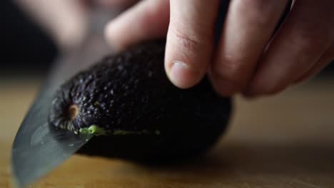 Hands-of-caucasian-male-rotating-avocado-on-wooden-chopping-board-while-slicing-it-in-half-with-sharp-chefs-knife,-SLOW-MOTION