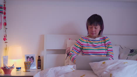 young-asian-malaysian-chinese-woman-sitting-in-bed-under-duvet-using-laptop-from-the-front