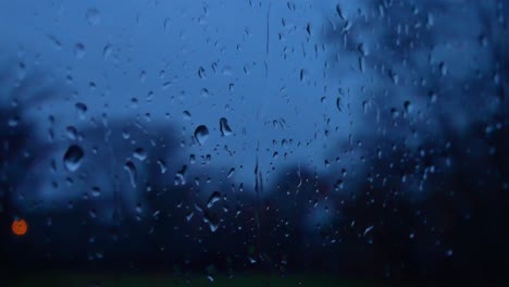 Rainy-storm-droplets-at-night,-moody-weather-fall-down-window-glass-closeup-bokeh