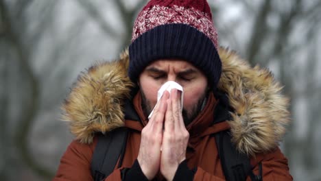 Bearded-Caucasian-male-places-white-paper-tissue-over-his-face-and-blows-his-nose-in-forest-during-cold-winter-day,-SLOW-MOTION
