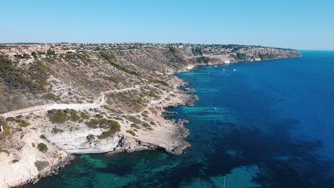 Aerial-drone-shot-in-4k-of-the-beautiful-rocky-coastline-of-the-balearic-island-Mallorca-with-clear-blue-and-turquoise-water-next-to-the-cliffs-with-boats-and-sailing-yachts-in-the-Mediterranean-Sea