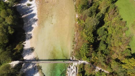 Top-view-drone-shot-of-tourist-walking-across-rope-bridge-in-alpine-environment,-clear-river-flows-under-bridge,-green-fields-and-trees-on-riverbeds,-sunny-summer-day