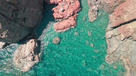 Blick-Von-Oben-Auf-Die-Roten-Felsen-Mit-Brechenden-Wellen-In-Einem-Wunderschönen-Blauen-Meer-Und-Tropischem-Wasser-An-Einem-Sonnigen-Tag-In-Sardinien,-Italien---Drohnenaufnahme-Aus-Der-Luft