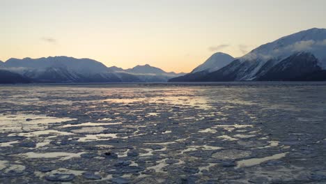 Dawn-at-large-open-icy-bay-surrounded-by-mountains-as-backdrop,-aerial-backwards