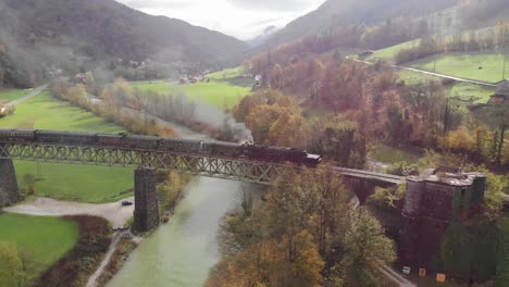 Old-steam-train-locomotive-with-wagons-drives-across-railway-bridge-in-countryside,-river-flows-below,-smoke-coming-from-engines,-overcast-late-summer-day