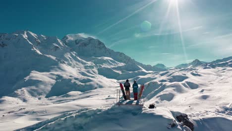 Snowboarders-on-incredible-mountaintop,-aerial-rising-rear-view,-mountain-reveal-backdrop