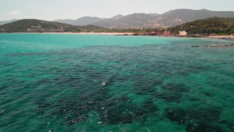 Fly-Over-Serene-Tropical-Ocean-With-Forested-Mountain-Ridges-At-Background-During-Summer-In-Sardinia,-Island-Of-Italy