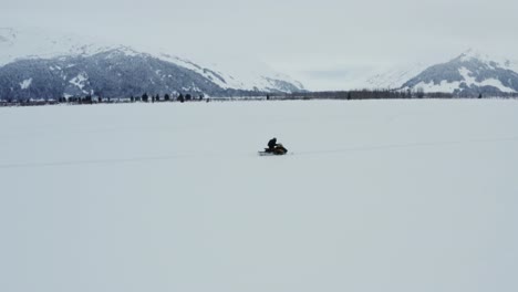 Man-on-snowmobile-traveling-on-snowy-field-with-mountains-in-background,-aerial