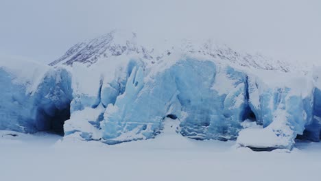 Vorderseite-Des-Gletschers-Mit-Blauem-Eis-Und-Mehreren-Höhlen,-Berge-Als-Hintergrund,-Antenne