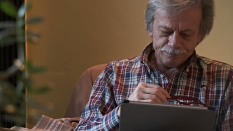 Front-View-Of-A-Man-Sitting-On-Couch-Using-Tablet-With-Gray-Haired,-Takes-Off-His-Glasses-And-Looks-In-Front---Slow-Motion-Shot