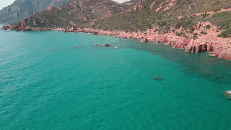 Drone-View-Near-The-Coast-Of-Red-Mountains-And-Green-Vegetation-In-Tropical-Blue-Sea-with-Waves-Crashing-on-the-Rocks-In-Sardinia,-Italy---Aerial-Shot