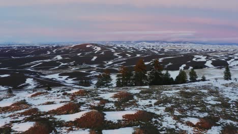 Flying-over-hill-to-reveal-a-farm-in-snow-covered-valley-during-soft-pink-sunset