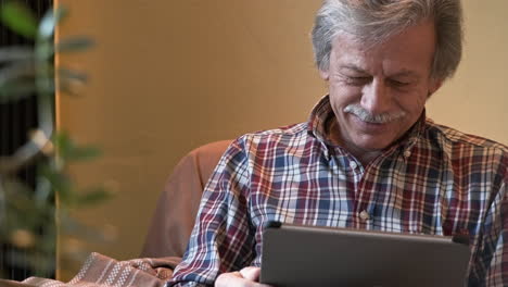 Senior-With-Gray-Haired-Looks-In-Front-And-Smile-While-Sitting-On-The-Couch-With-His-Tablet---Slow-Motion-Shot