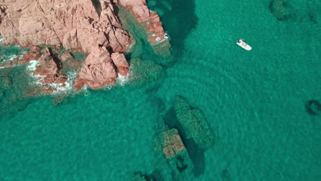 Static-View-From-Above-Of-The-Transparent-And-Crystalline-Sea-Where-The-Waves-Crash-Against-The-Red-Rocks-On-A-Boat-With-Tourists-Swimming-Under-the-Sun-In-Sardinia,-Italy---Aerial-Shot