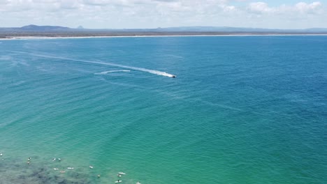 Drone-descending-on-a-peninsula-beach,-boat-and-surfers-in-the-background