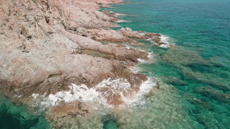 Aerial-View-Of-The-Foam-Waves-Crashing-Against-The-Cliff-Above-The-Transparent-Sea-On-A-Sunny-Day-In-August-During-Summer-Holiday-In-Sardinia,-Italy---Drone-Wide-Shot-in-Slow-Motion