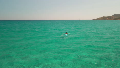 Rear-View-Of-Man-On-Kayak-Sailing-Alone-In-The-Clear-Blue-Sea-Under-The-August-Sun-In-Sardinia,-Italy---aerial-drone-shot
