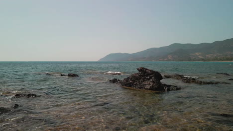 Meereswellen,-Die-Während-Des-Sommernachmittags-In-Sardinien,-Italien,-Auf-Die-Felsen-Mit-Bergen-Im-Hintergrund-Schlagen---Luftdrohnenaufnahme