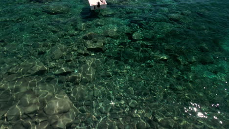 Filmischer-Blick-Vom-Kieselstrand-über-Das-Transparente-Meer,-Das-An-Einem-Sonnigen-Tag-In-Sardinien,-Italien,-über-Festgemachte-Boote-Fährt---Luftdrohnenaufnahme