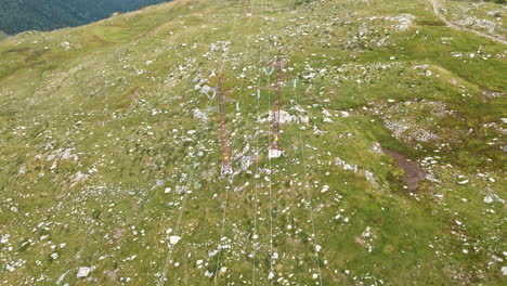 Hochspannungskabel-Mit-Masten-Auf-Felsiger-Grüner-Wiese-Der-Alpen-Im-Norden-Italiens---Luftdrohnenaufnahme