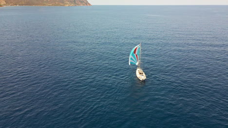 Sailboat-With-Colorful-Sail-Cruising-In-Blue-Sea-Towards-The-Coast-In-The-Background-In-Sardinia,-Italy---aerial-drone-shot