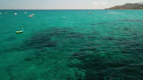 Blaugrünes-Wasser-Des-Mittelmeeres-Mit-Festgemachten-Booten-Und-Bergen-Im-Hintergrund-Während-Des-Sommers-In-Sardinien,-Italien---Luftrückzug