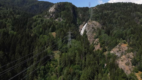 Elektrische-Kabel-Führen-Durch-Den-Grünen-Wald-In-Den-Schneebedeckten-Bergen-Mit-Kaskaden-Im-Hintergrund-In-Italien