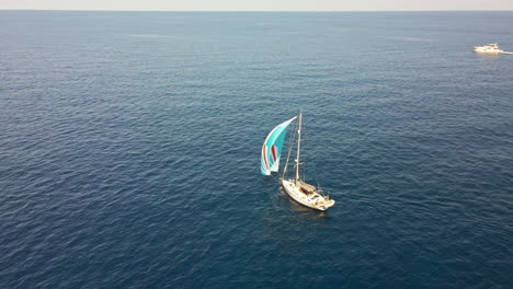 Segelboot-Mit-Buntem-Segel,-Das-Im-Blauen-Meer-Während-Des-Sonnenuntergangs-Des-Sommers-In-Sardinien,-Italien-Kreuzt---Luftdrohne-Weit-Geschossen