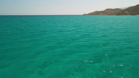 Fliegen-über-Das-Tropische-Blaugrüne-Wasser-Des-Mittelmeers-Mit-Segelbooten-Und-Küste-Im-Hintergrund-Sardiniens,-Italien---Luftdrohnenaufnahme