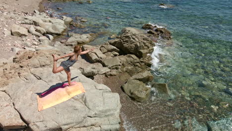 Beautiful-Woman-Maintains-Yoga-Position-On-A-Rock-By-The-Sea-In-Sardinia,-Italy---Aerial-drone-zoom-in-shot-in-slow-motion