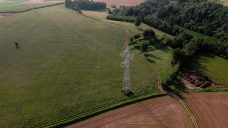 Antenne-Des-Stromturms-Und-Der-Stromleitungen-über-Dem-Grünen-Landwirtschaftsfeld-An-Einem-Sonnigen-Sommerabend,-Drohnenaufnahme-Im-Orbit