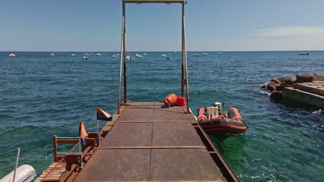 Fliegen-über-Den-Pier-Unter-Der-Sonne-Mit-Festgemachtem-Schlauchboot-Und-Wellen-Des-Blauen-Meeres-Von-Sardinien,-Italien---Luftdrohne-In-Zeitlupe-Aufgenommen